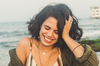 A person is laughing joyfully while holding their head with one hand. They are wearing a striped dress with an open shirt over it, several necklaces, and rings. The background features the sea with soft waves and a structure on the horizon.