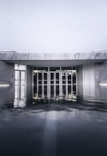 An architectural structure with a modern design is partially submerged in water, with reflections creating a mirror-like effect on the surface. The building appears to be made of gray stone with prominent geometric lines, and there are large glass doors at the entrance.