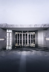 An architectural structure with a modern design is partially submerged in water, with reflections creating a mirror-like effect on the surface. The building appears to be made of gray stone with prominent geometric lines, and there are large glass doors at the entrance.