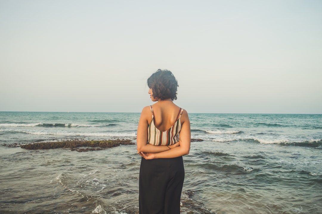 woman in striped spaghetti strap top and black pants facing ocean