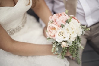 A close-up of a bride holding a bouquet of flowers, featuring pink and white blooms with green foliage. The bride is wearing a white lace dress with a decorative waistband, and there is another person in the background, dressed in a light-colored shirt and beige pants.