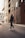 A person wearing a dark hoodie and stylish sneakers walks down an urban street. Tall modern buildings with glass windows line the street, reflecting city life. A vehicle is visible on the left, creating a sense of movement and energy.