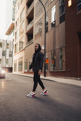 A person wearing a dark hoodie and stylish sneakers walks down an urban street. Tall modern buildings with glass windows line the street, reflecting city life. A vehicle is visible on the left, creating a sense of movement and energy.