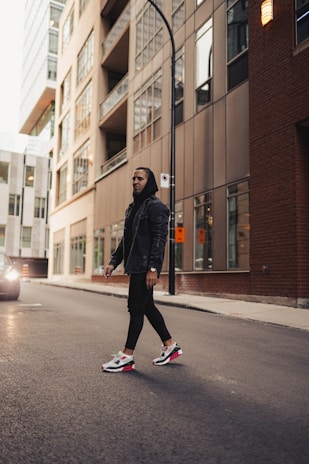 A person wearing a dark hoodie and stylish sneakers walks down an urban street. Tall modern buildings with glass windows line the street, reflecting city life. A vehicle is visible on the left, creating a sense of movement and energy.