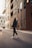 A person wearing a dark hoodie and stylish sneakers walks down an urban street. Tall modern buildings with glass windows line the street, reflecting city life. A vehicle is visible on the left, creating a sense of movement and energy.