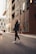 A person wearing a dark hoodie and stylish sneakers walks down an urban street. Tall modern buildings with glass windows line the street, reflecting city life. A vehicle is visible on the left, creating a sense of movement and energy.