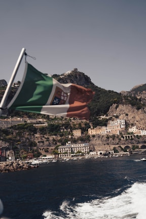 A flag with green, white, and red sections waves in the foreground. In the background, a coastal town with pastel-colored buildings is nestled against steep, rocky cliffs. The sea below is a deep blue with white waves, and a few boats are visible near the shore.