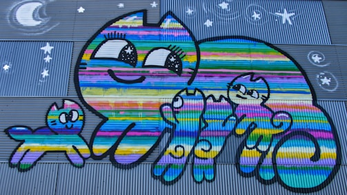 A colorful mural features a large cartoon-style cat and three smaller cats, all adorned with bright, multicolored stripes. The cats are smiling and appear cheerful, with large, expressive eyes. The background includes a corrugated metal surface with painted stars and a crescent moon, conveying a night sky theme.