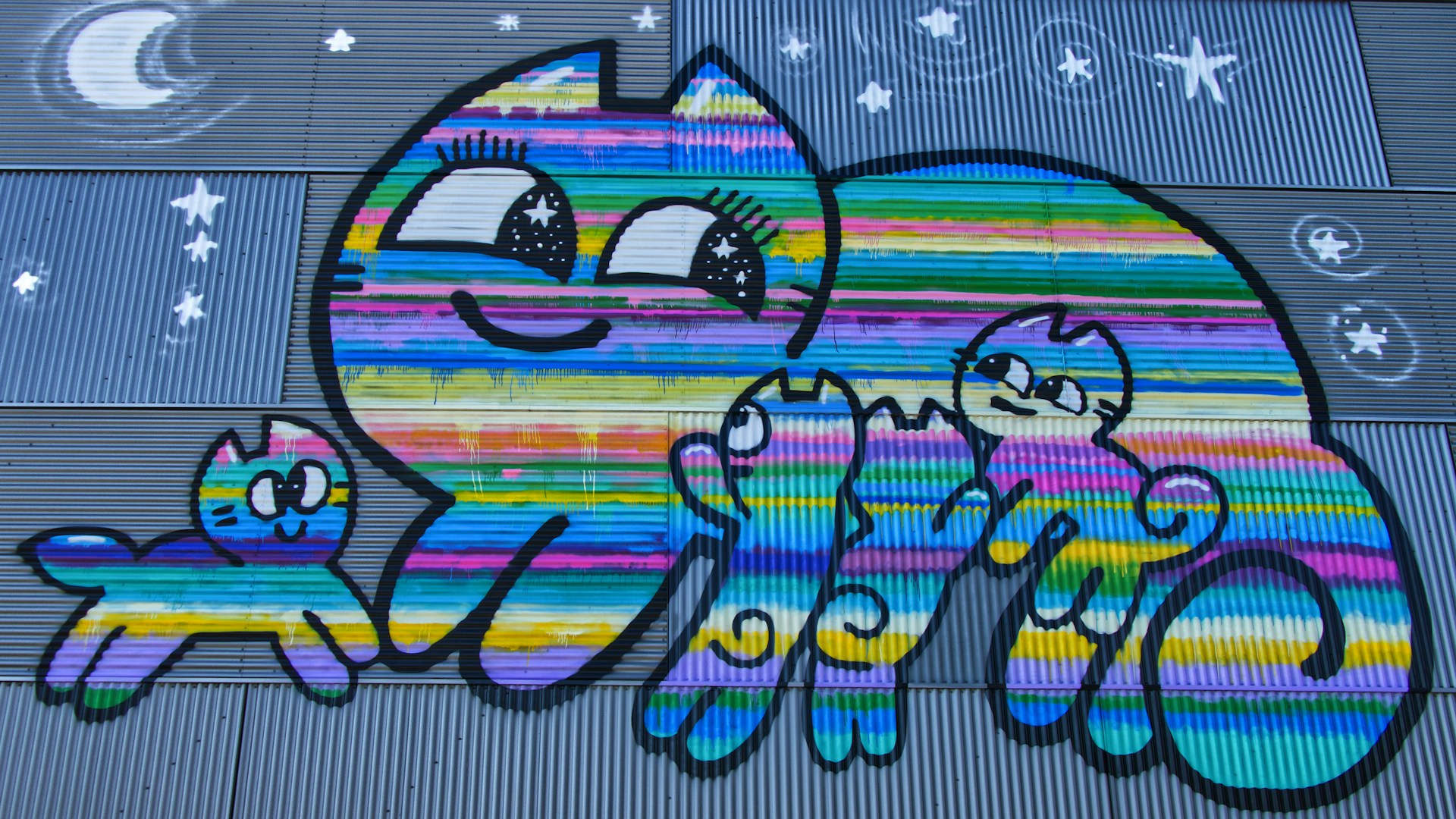 A colorful mural features a large cartoon-style cat and three smaller cats, all adorned with bright, multicolored stripes. The cats are smiling and appear cheerful, with large, expressive eyes. The background includes a corrugated metal surface with painted stars and a crescent moon, conveying a night sky theme.