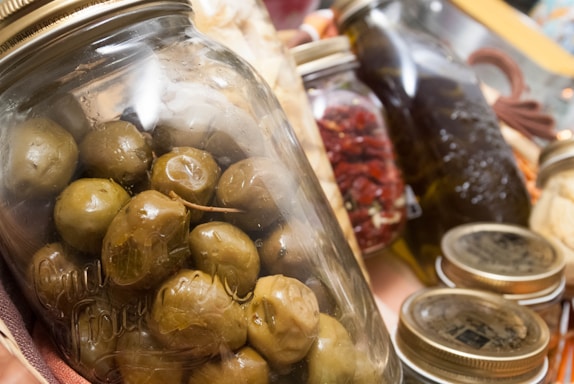 A close-up view of several jars containing pickled and preserved foods, including green olives and sundried tomatoes. The jars are arranged on a surface and include metal lids and visible condensation.