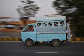A light blue van is speeding on a road with a blurred background, indicating motion. The vehicle appears to have several passengers inside, with one person sitting at the back, holding onto the exterior. The scene suggests a busy, possibly urban setting.