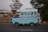 A light blue van is speeding on a road with a blurred background, indicating motion. The vehicle appears to have several passengers inside, with one person sitting at the back, holding onto the exterior. The scene suggests a busy, possibly urban setting.