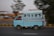 A light blue van is speeding on a road with a blurred background, indicating motion. The vehicle appears to have several passengers inside, with one person sitting at the back, holding onto the exterior. The scene suggests a busy, possibly urban setting.