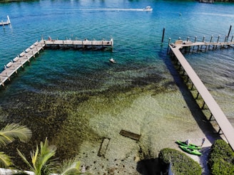 A scenic view of a clear, emerald coastline with wooden piers extending over the water. Two people are standing near bright green kayaks on the shore. The water reveals the textures and patterns of the seabed below. A speedboat is moving swiftly across the water in the background.