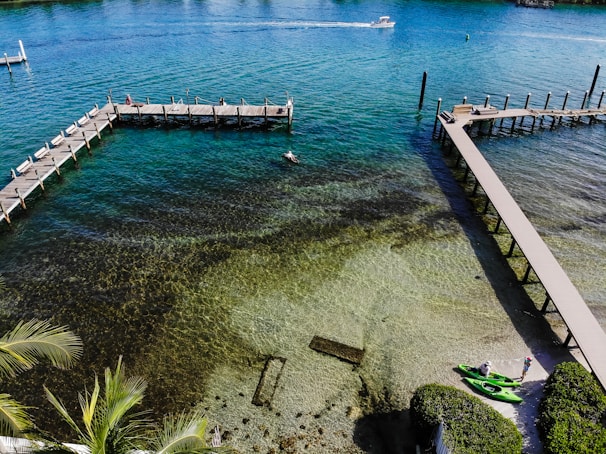 A scenic view of a clear, emerald coastline with wooden piers extending over the water. Two people are standing near bright green kayaks on the shore. The water reveals the textures and patterns of the seabed below. A speedboat is moving swiftly across the water in the background.