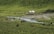 A serene pastoral landscape features a group of horses grazing on lush green grass near a flowing stream. In the background, two white yurts are situated, surrounded by small rocks and gently sloping hills covered in vegetation.