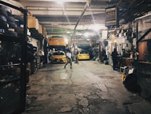 A dimly lit auto repair garage with two yellow cars, one on a lift and another below it. Various car parts, tools, and equipment are scattered around. A person is working at a desk on the right, while another person walks in the center. Shelves on the left are filled with tires and other items.