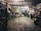 A dimly lit auto repair garage with two yellow cars, one on a lift and another below it. Various car parts, tools, and equipment are scattered around. A person is working at a desk on the right, while another person walks in the center. Shelves on the left are filled with tires and other items.