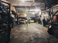 A dimly lit auto repair garage with two yellow cars, one on a lift and another below it. Various car parts, tools, and equipment are scattered around. A person is working at a desk on the right, while another person walks in the center. Shelves on the left are filled with tires and other items.