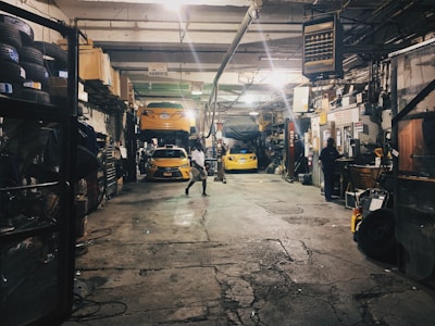 A dimly lit auto repair garage with two yellow cars, one on a lift and another below it. Various car parts, tools, and equipment are scattered around. A person is working at a desk on the right, while another person walks in the center. Shelves on the left are filled with tires and other items.