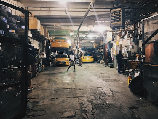A dimly lit auto repair garage with two yellow cars, one on a lift and another below it. Various car parts, tools, and equipment are scattered around. A person is working at a desk on the right, while another person walks in the center. Shelves on the left are filled with tires and other items.