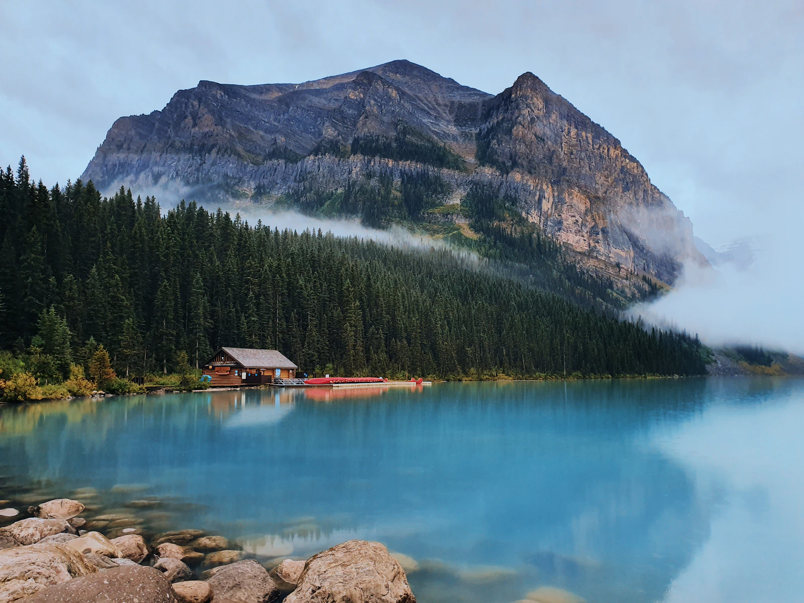 A calm Canadian mountain lake mirroring a rugged ridge and evergreen forest under a misty overcast sky