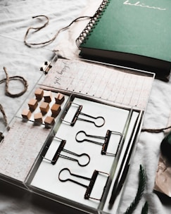 An organized workspace setup featuring a green spiral notebook, a tray with large binder clips, a pen, wooden cubes, and note cards. Twine and a piece of pine or spruce are also present, giving a rustic and natural feel to the arrangement.