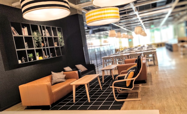 A modern, cozy lounge area with a variety of seating options including two couches and a chair arranged around a light wood coffee table. The floor is covered with a black checkered rug, and the walls have a black shelving unit decorated with books and green plants. The room is well-lit by large, hanging light fixtures with black and white, and yellow-striped patterns. The background features a dining area with multiple tables and chairs, under additional hanging light fixtures.