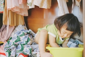 A young child is sitting inside a wardrobe among hanging clothes and a pile of colorful fabric. The child is wearing a light green and white shirt and is focused on reaching into a yellow plastic basket, as though playing or searching for something. The scene is warmly lit, providing a cozy and playful atmosphere.