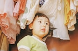 A young child with short dark hair gazes upward, surrounded by various pieces of clothing hanging above. The clothes have a variety of soft, pastel colors such as pink, white, yellow, and green, adding a cozy and playful atmosphere to the scene.