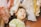 A young child with short dark hair gazes upward, surrounded by various pieces of clothing hanging above. The clothes have a variety of soft, pastel colors such as pink, white, yellow, and green, adding a cozy and playful atmosphere to the scene.