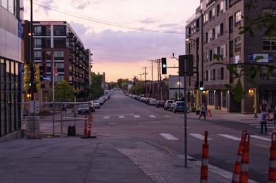 A city street scene at sunset, lined with parked cars on both sides and modern apartment buildings. Traffic lights are visible, and a couple of people are walking across the street. A sign on a building indicates 'Now Leasing.' The sky is partly cloudy with hints of pink and orange from the setting sun.