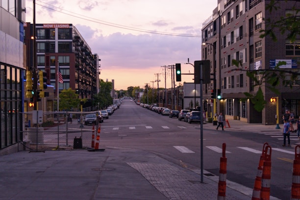 A city street scene at sunset, lined with parked cars on both sides and modern apartment buildings. Traffic lights are visible, and a couple of people are walking across the street. A sign on a building indicates 'Now Leasing.' The sky is partly cloudy with hints of pink and orange from the setting sun.