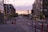 A city street scene at sunset, lined with parked cars on both sides and modern apartment buildings. Traffic lights are visible, and a couple of people are walking across the street. A sign on a building indicates 'Now Leasing.' The sky is partly cloudy with hints of pink and orange from the setting sun.