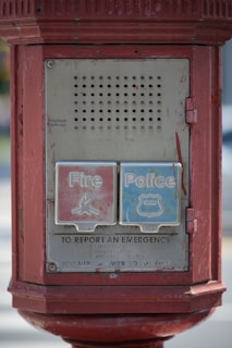 A vintage emergency call box with two large buttons labeled 'Fire' and 'Police'. The box is rectangular, made of metal, and painted red. It features instructions for reporting emergencies below the buttons and displays visible signs of age with worn-out paint.