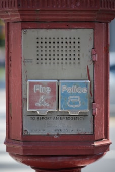 A vintage emergency call box with two large buttons labeled 'Fire' and 'Police'. The box is rectangular, made of metal, and painted red. It features instructions for reporting emergencies below the buttons and displays visible signs of age with worn-out paint.