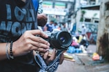 A person holds a vintage camera with a strap, displaying an interest in photography. The background is an urban street scene, with blurred people and vibrant market colors, suggesting a busy, lively atmosphere.
