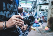 A person holds a vintage camera with a strap, displaying an interest in photography. The background is an urban street scene, with blurred people and vibrant market colors, suggesting a busy, lively atmosphere.