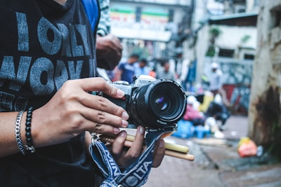 A person holds a vintage camera with a strap, displaying an interest in photography. The background is an urban street scene, with blurred people and vibrant market colors, suggesting a busy, lively atmosphere.