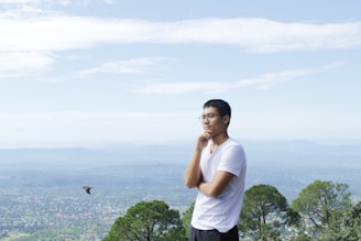 A man wearing glasses and a white t-shirt stands thoughtfully with his hand on his chin. He is overlooking a vast, expansive landscape featuring distant hills and scattered greenery. A bird is flying nearby in the clear, blue sky.