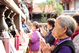 A group of elderly people is standing in a line, hands clasped in prayer or contemplation, in what appears to be a temple or shrine setting. They are surrounded by stone railings and various religious or cultural objects. The focus is on a woman in the foreground with gray hair wearing a black vest over a purple shirt.