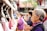 A group of elderly people is standing in a line, hands clasped in prayer or contemplation, in what appears to be a temple or shrine setting. They are surrounded by stone railings and various religious or cultural objects. The focus is on a woman in the foreground with gray hair wearing a black vest over a purple shirt.