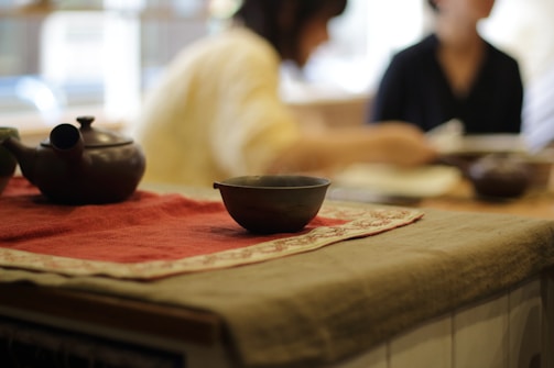 A table is set with a dark ceramic teapot and bowl placed on a richly colored red cloth. The background is softly blurred, showing people seated, suggesting a peaceful and intimate gathering or tea service.