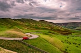 A winding road travels through lush green hills with a vibrant red van traveling along it. The landscape is expansive, with undulating terrain and a cloudy sky overhead, adding a moody feel to the scene.