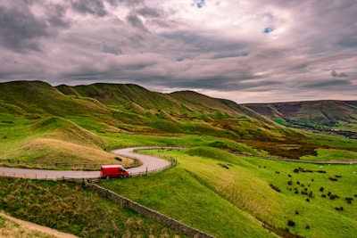 A winding road travels through lush green hills with a vibrant red van traveling along it. The landscape is expansive, with undulating terrain and a cloudy sky overhead, adding a moody feel to the scene.