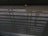 A reflective surface captures the pattern of shadows and a structural lattice above. In the background, a small object, possibly a crumpled paper or piece of trash, is situated on the floor. The shadows create a striped effect on the otherwise neutral ground, and the reflection presents layered dimensions.