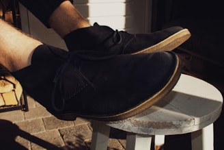 A close-up shot of a pair of black suede shoes with light brown soles, worn by a person. The shoes are resting on a white wooden stool, placed on a stone-tiled surface. The background includes a shadowy area with some wood storage and a partially visible door.