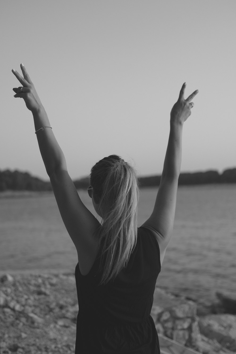 grayscale photo of woman wearing black tank top walking on seashore while doing peace hand sign