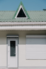 A building facade with a white door and a closed window shutter. Above these, there is a green tiled roof featuring a triangular dormer window with a black frame.