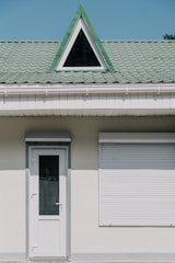 A building facade with a white door and a closed window shutter. Above these, there is a green tiled roof featuring a triangular dormer window with a black frame.